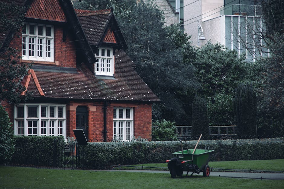 View of a row of Victorian-style residential houses in Swiss Cottage, featuring ornate white wooden balcony railings, large bay windows, and decorative gables with red brick facades. The houses are situated on a slightly elevated street with a brick retaining wall, landscaped with small bushes and trees. In the foreground, there are several black wheelie bins lined along the pavement, and the street appears clean and well-maintained under bright daylight with a clear blue sky. This image exemplifies residential architecture in NW3 area, highlighting the importance of exterior surface cleaning and maintenance, as offered by Swiss Cottage Cleaners in their domestic cleaning services.