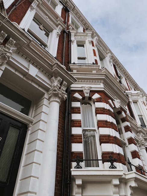 Photograph of an ornate residential building facade featuring red brickwork combined with white decorative elements. The structure includes large arched windows with white frames and sills, some adorned with small wrought iron balconies. Decorative white columns with Corinthian capitals flank the windows, adding architectural detail. The exterior showcases a mix of brick and stucco, with intricate cornices and decorative mouldings under a partly cloudy sky. The building is clean and well-maintained, reflecting high-quality surface cleaning consistent with services offered by Swiss Cottage Cleaners for end of tenancy cleaning on Adelaide Road tenants in Swiss Cottage. Natural daylight illuminates the façade, highlighting the detailed craftsmanship.