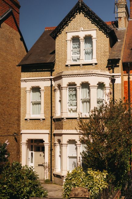 Photograph of an ornate residential building facade featuring red brickwork combined with white decorative elements. The structure includes large arched windows with white frames and sills, some adorned with small wrought iron balconies. Decorative white columns with Corinthian capitals flank the windows, adding architectural detail. The exterior showcases a mix of brick and stucco, with intricate cornices and decorative mouldings under a partly cloudy sky. The building is clean and well-maintained, reflecting high-quality surface cleaning consistent with services offered by Swiss Cottage Cleaners for end of tenancy cleaning on Adelaide Road tenants in Swiss Cottage. Natural daylight illuminates the façade, highlighting the detailed craftsmanship.