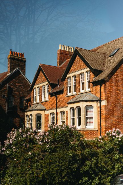 An exterior view of a traditional red brick residential building with multiple pitched roofs and white-framed windows, situated amidst a garden with flowering bushes. The building's surfaces appear cl