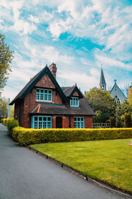 An exterior view of a traditional red brick residential building with multiple pitched roofs and white-framed windows, situated amidst a garden with flowering bushes. The building's surfaces appear cl
